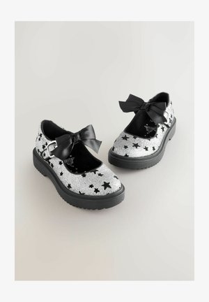 Chaussures Mary Jane en paillettes argentées avec des motifs d'étoiles noires, des accents noirs, une semelle en caoutchouc plate et un grand nœud en ruban noir.