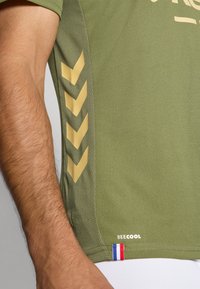 Olivenfarvet atletisk skjorte i åndbart stof, med gyldne chevron-detaljer på siden og logo med tricolore detalje i kanten.