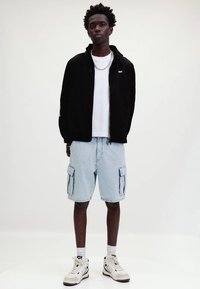 Sort zip-up jakke, hvid t-shirt, lyseblå cargo shorts med sidelommer og cremede sneakers. Model står foran en neutral baggrund.