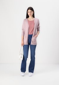 Cardigan lavorato a maglia rosa chiaro con maniche larghe, abbinato a una camicia rosa e jeans a zampa d'elefante blu. Il modello tiene una piccola borsa bianca.