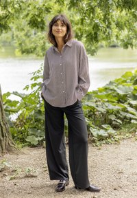 Striped button-up shirt in light and dark colors, paired with black wide-leg pants and shiny loafers, standing by a water feature surrounded by greenery.