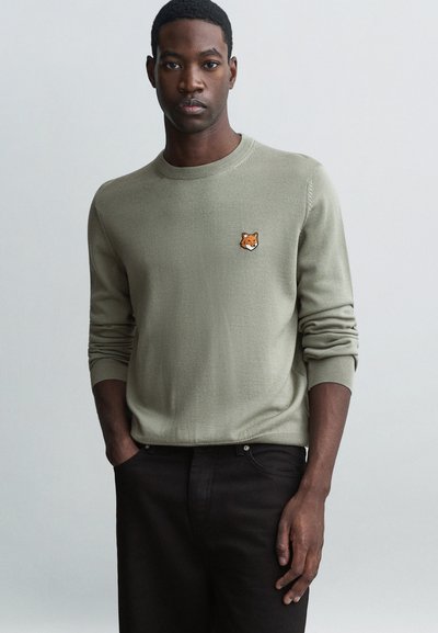 Pull en maille vert olive clair avec un col rond, orné d'une petite broderie de tête de renard orange sur la poitrine. Assorti avec un pantalon sombre.