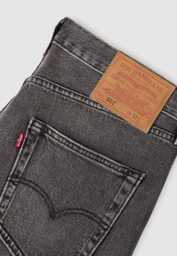 Διπλωμένο γκρι τζιν Levi's 501 που δείχνει την πίσω τσέπη με κόκκινη ετικέτα Levi's και ταμπά δερμάτινο έμπλαστρο με λεπτομέρειες μάρκας και μεγέθους.