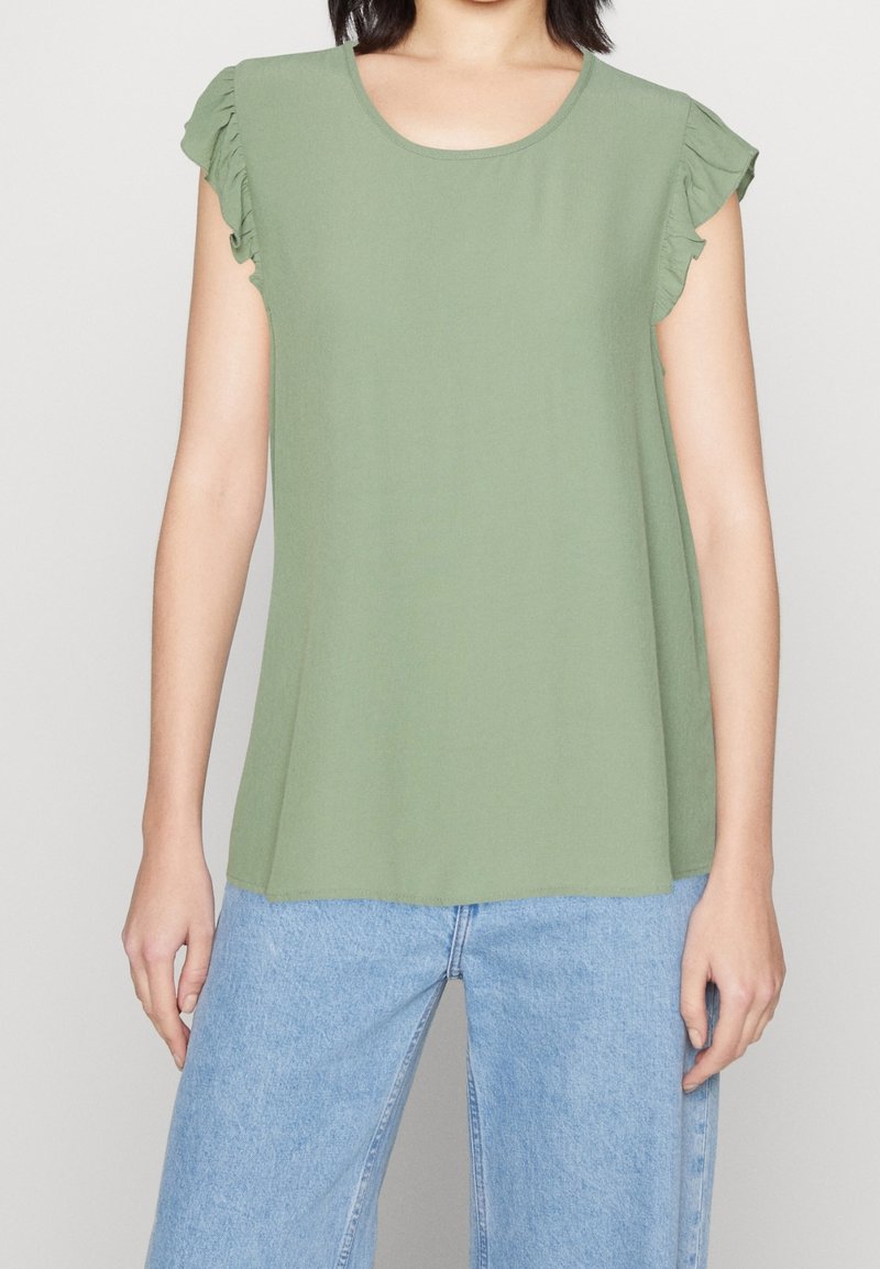 Oliwkowy bezrękawnik z falbaniastymi akcentami na ramionach, okrągłym dekoltem i luźnym krojem, połączony z jasnoniebieskimi spodniami typu wide-leg.