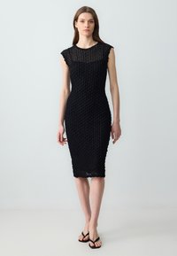 Robe noire ajustée avec un design texturé et à motifs, manches courtes et longueur mi-mollet. Les chaussures comprennent des simples tongs noires.