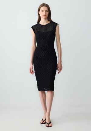 Robe noire ajustée avec un design texturé et à motifs, manches courtes et longueur mi-mollet. Les chaussures comprennent des simples tongs noires.