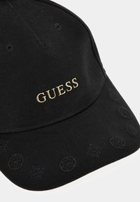 Sort kasket lavet af bomuld med en krum skygge. Har det gyldne "GUESS" logo og sorte broderede cirkulære mønstre langs skyggen.