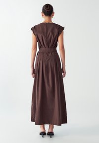 Žena s vlasy v drdolu, oblečená v hnědé midi šaty bez rukávů s sklady a páskem, stojící na černých vysokých podpatcích na bílém pozadí.