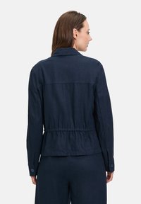 Granatowa kurtka z teksturowanego materiału, z zaciśnięciem w talii, guzikami na mankietach i kołnierzem, widziana z tyłu.