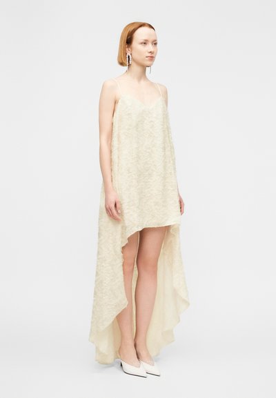 Εκρού, ασύμμετρο φόρεμα με υφασμάτινη υφή. Διαθέτει λεπτές τιράντες και είναι μακρύτερο στο πίσω μέρος. Συνδυάζεται με λευκά μυτερά παπούτσια.