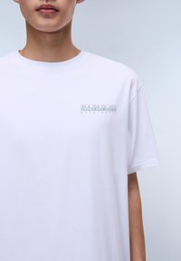 Camiseta blanca de manga corta con corte holgado, que presenta un logo impreso en verde y gris en el área del pecho izquierdo. Textura de algodón suave.