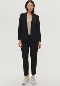 Zwarte getailleerde blazer met één knoop, gecombineerd met bijpassende zwarte taps toelopende broeken en een beige top. Afgemaakt met witte sneakers.