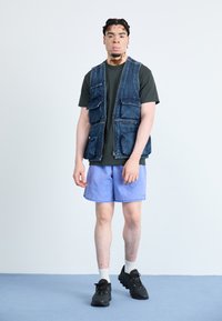 Denim prsluk s više prednjih džepova, u kombinaciji s tamnom majicom i svijetloplavim šortsevima. Crne atletske cipele i bijele čarape upotpunjuju izgled.