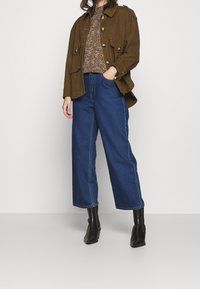 Olive green utility jacket with button details, paired with a patterned brown top and wide-leg blue jeans. Black ankle boots complete the outfit.