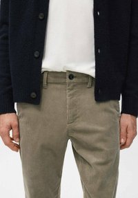 Man draagt beige corduroy broek, wit overhemd en zwarte cardigan met knopen, handen ontspannen langs het lichaam.