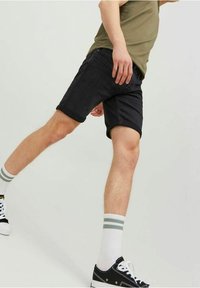 Persona que lleva pantalones cortos de mezclilla negros, calcetines blancos con rayas verdes, zapatillas negras y una camiseta verde oliva, levantando una pierna.