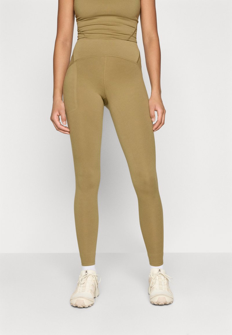 Olivno zelene legging hlače visokega pasu iz elastičnega materiala, brezšivnega dizajna in stranskih žepov, v kombinaciji s bež športnimi čevlji.