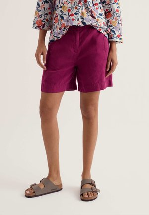 Persoană care poartă pantaloni scurți magenta, un top cu mâneci lungi cu flori și sandale gri în stil Birkenstock, stând în fața unui fundal simplu.