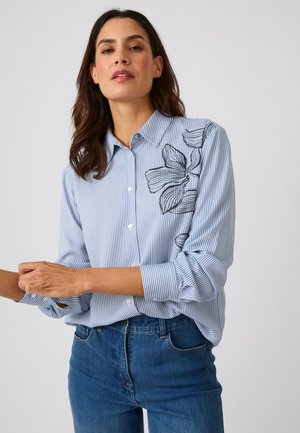 Frau trägt ein blau-weiß gestreiftes Hemd mit Knöpfen und schwarzer Blumenstickerei auf der Schulter, kombiniert mit blauen Jeans.