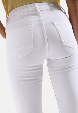 Nahaufnahme einer Person, die eng anliegende weiße Jeans mit Gesäßtaschen und einem kleinen Markenschild am Bund trägt, kombiniert mit einem gelben Oberteil.