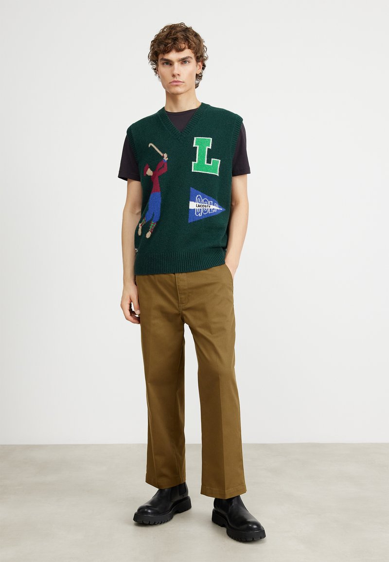 Gilet lavorato a maglia verde con grafica ricamata di un golfista colorato, loghi "L" e "Lacoste". Indossato sopra una maglietta nera. Pantaloni beige e stivali neri.