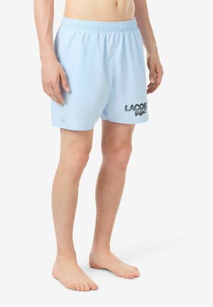 Hombre con bañador corto Lacoste azul claro, descalzo sobre un fondo blanco.