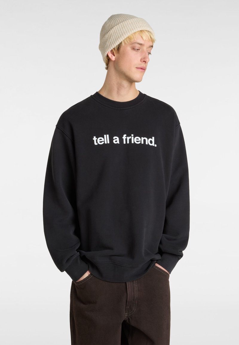 Sort sweatshirt med hvid tekst "tell a friend." Klassisk crew neck design, afslappet pasform og blødt stof. Brune bukser fremhævet.