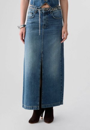 Donna che indossa una lunga gonna di denim con uno spacco frontale, abbinata a stivaletti neri e una cintura a catena argentata.