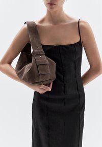 Brown suede handbag with a wide strap and a folded design, featuring a snap closure and contrasting white stitching. Worn with a black dress.