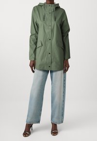 Grön vattentät jacka med huva, tryckknappar, två framfickor och en avslappnad passform, kombinerad med ljusblå jeans med vida ben och svarta sandaler.