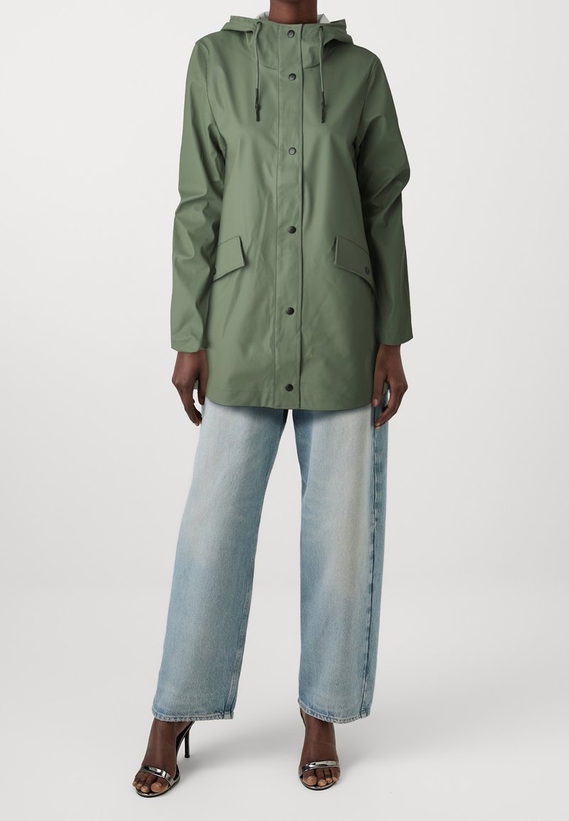 Grön vattentät jacka med huva, tryckknappar, två framfickor och en avslappnad passform, kombinerad med ljusblå jeans med vida ben och svarta sandaler.