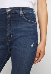 Jeans de mezclilla azul oscuro con una cintura media, diseño clásico de cinco bolsillos, costuras visibles y un ligero desgaste sutil en el muslo.