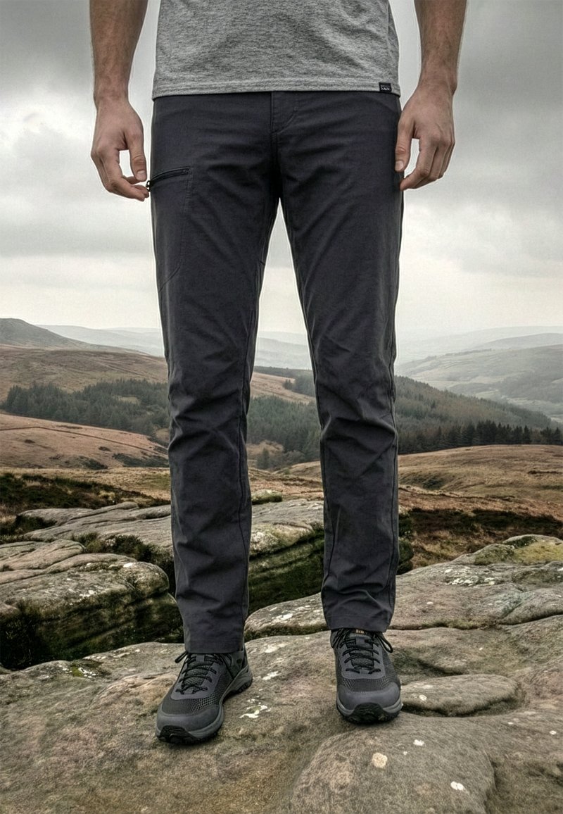 Muž stojaci na skalnatom teréne, oblečený v tmavé turistické nohavice a čierne trailové topánky s hmlistou, kopcovitou krajinou v pozadí.