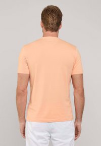 Camiseta de manga corta color melocotón, hecha de material suave, con cuello redondo y un diseño liso. Vista trasera con un branding mínimo cerca del cuello.