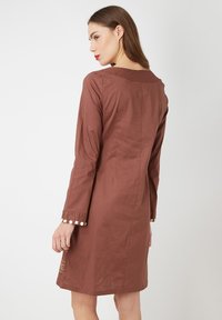 Vestido marrón con mangas largas, que incluye acentos de botones blancos en los puños y un diseño simple y recto. Textura de tela suave.