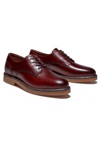 Paire de chaussures habillées marron foncé en cuir verni, à lacets et semelles en caoutchouc, présentées en angle pour montrer les côtés extérieur et avant sur fond blanc.