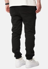 Black jogger pants with a slim fit, featuring cuffed ankles and a back pocket, paired with white sneakers. Smooth fabric texture.