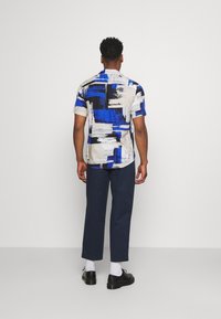 Kortärmad skjorta med ett abstrakt mönster i blått, svart och beige, med en lätt rundad nederkant. Matchad med marinblå byxor och svarta skor.