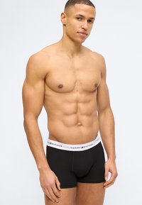 Μυώδης άντρας με κοντά μαλλιά που φοράει μαύρα εσώρουχα Tommy Hilfiger με λευκή μέση, στέκεται μπροστά σε απλό ανοιχτό φόντο.