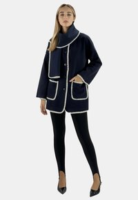 Abrigo de mezcla de lana en color navy con ribete blanco, grandes bolsillos, cierre de botones y un cuello ancho, combinado con leggings negros y tacones de punta.