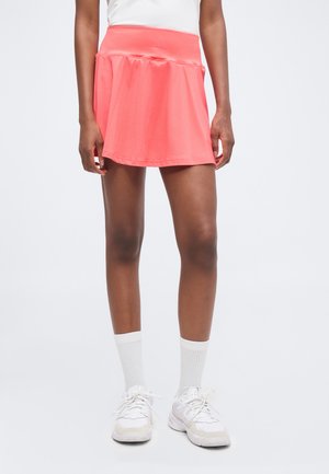 Weibliche Beine mit einem kurzen rosa Rock, weißen Kniestrümpfen und weißen Sportschuhen vor einem schlichten hellen Hintergrund.