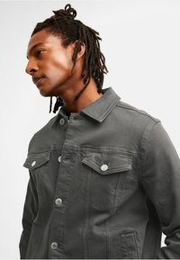 Grå denimjacka med krage, två framfickor med tryckknappar och knäppning fram, visar en texturerad tygytan.