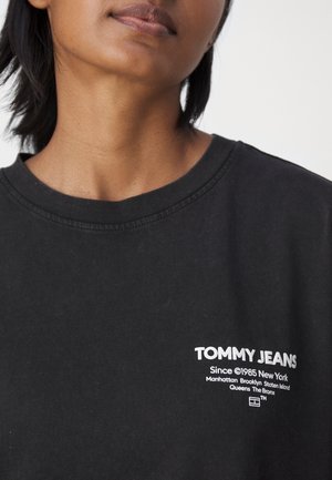Osoba nosząca czarną koszulkę Tommy Jeans z białym tekstem wymieniającym dzielnice Nowego Jorku na piersi.