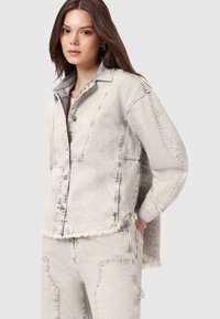 Jasnoszara dżinsowa kurtka z postrzępionym dołem, zapięcie na guziki i kontrastowe przeszycia, zestawiona z pasującymi jasnoszarymi spodniami dżinsowymi.