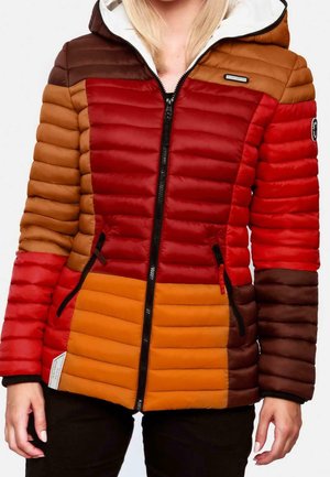 Femme portant une doudoune zippée à blocs de couleur rouge, orange et marron avec une capuche blanche, debout les mains détendues.