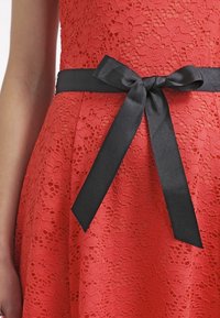 Robe en dentelle rouge avec un motif texturé et une taille cintrée. Une ceinture en satin noir est nouée en nœud au centre.