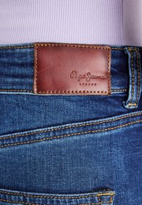 Sinised teksapüksid kollase õmblusega ja pruun nahast silt, millel on "Pepe Jeans London" vöökohtade all helelilla soonikkoes pluusi all.