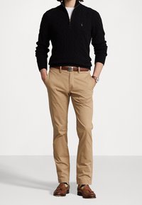 Schwarzer Kabelstrickpullover mit Viertelreißverschluss, beige Chinos, brauer Ledergürtel und braune Slipper. Detaillierte Nähte an der Kleidung.