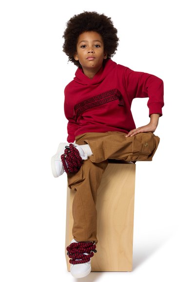 Felpa rossa con dettagli a strisce testurizzati, pantaloni cargo beige e sneaker bianche con accenti a motivo rosso e nero; seduto su un blocco chiaro.
