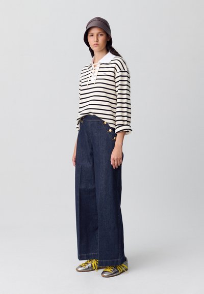 Camisa de mangas largas a rayas con cuello atado, combinada con pantalones de mezclilla oscuros de pierna ancha con botones dorados y zapatillas metálicas con cordones amarillos.